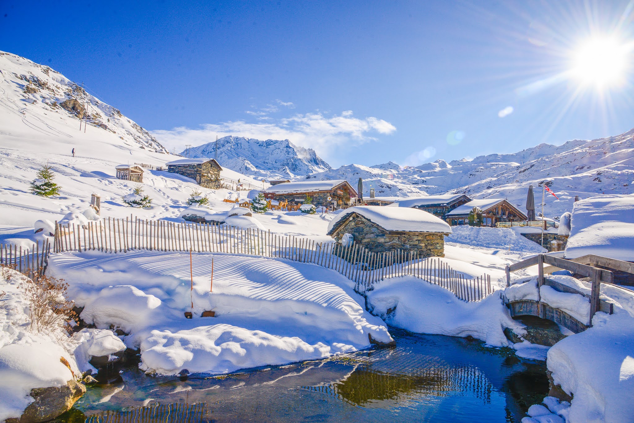 Bassin et petit pont en bois devant les chalets en pierre de Chez Pépé Nicolas, entourés de neige sous un soleil hivernal