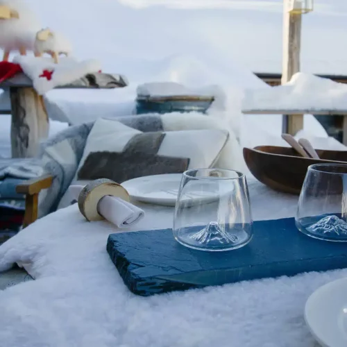 Verres à relief de montagne sur un plateau bleu, avec vaisselle et coussin à motif de croix dans un décor enneigé chez Pépé Nicolas.